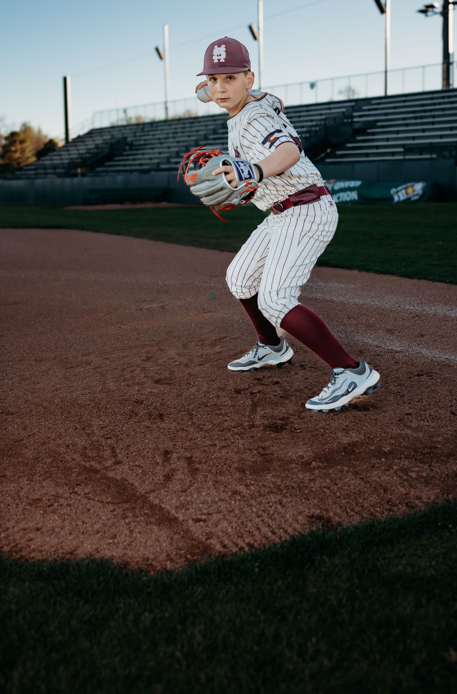 Young baseball player in a maroon cap and pinstriped uniform crouches in a fielding stance on the infield dirt, glove ready.