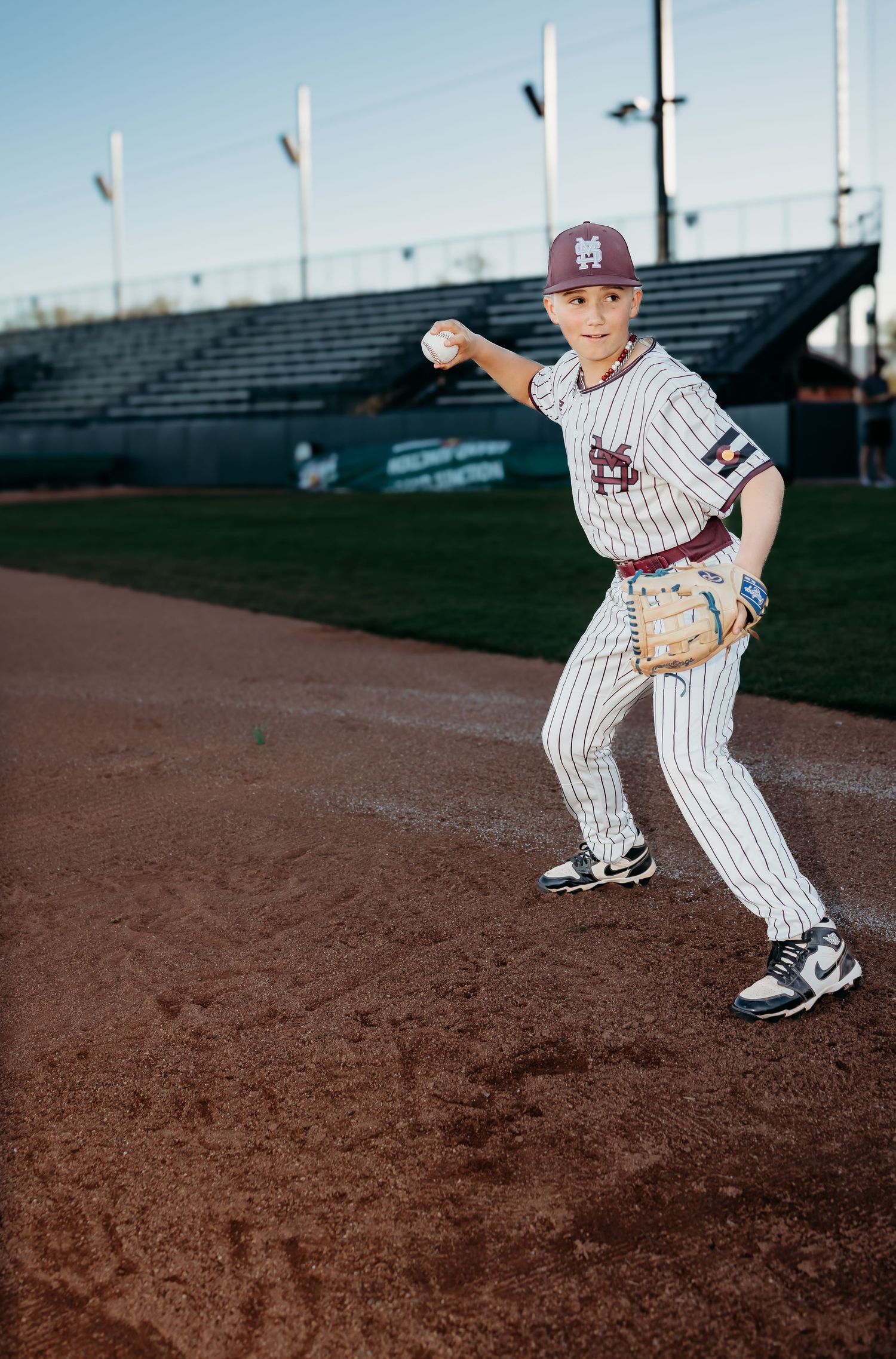 Youth baseball player in a pinstriped uniform and maroon cap winds up to throw a baseball on a dirt infield with stadium seating behind.