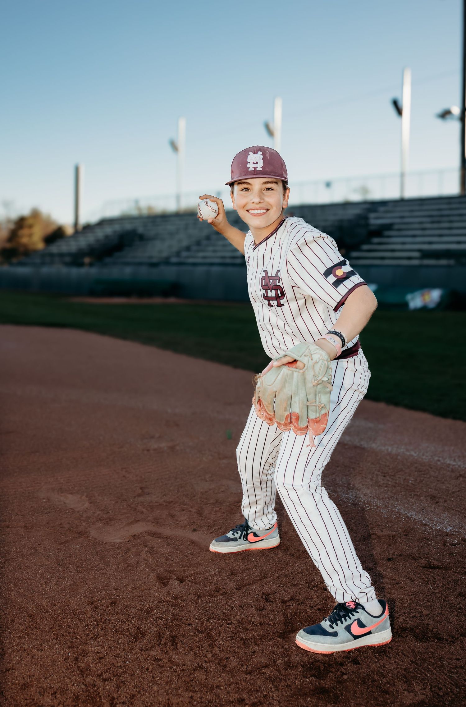 Smiling young baseball player in a maroon cap and pinstriped uniform holds a ball and glove, posing on the infield at sunset.