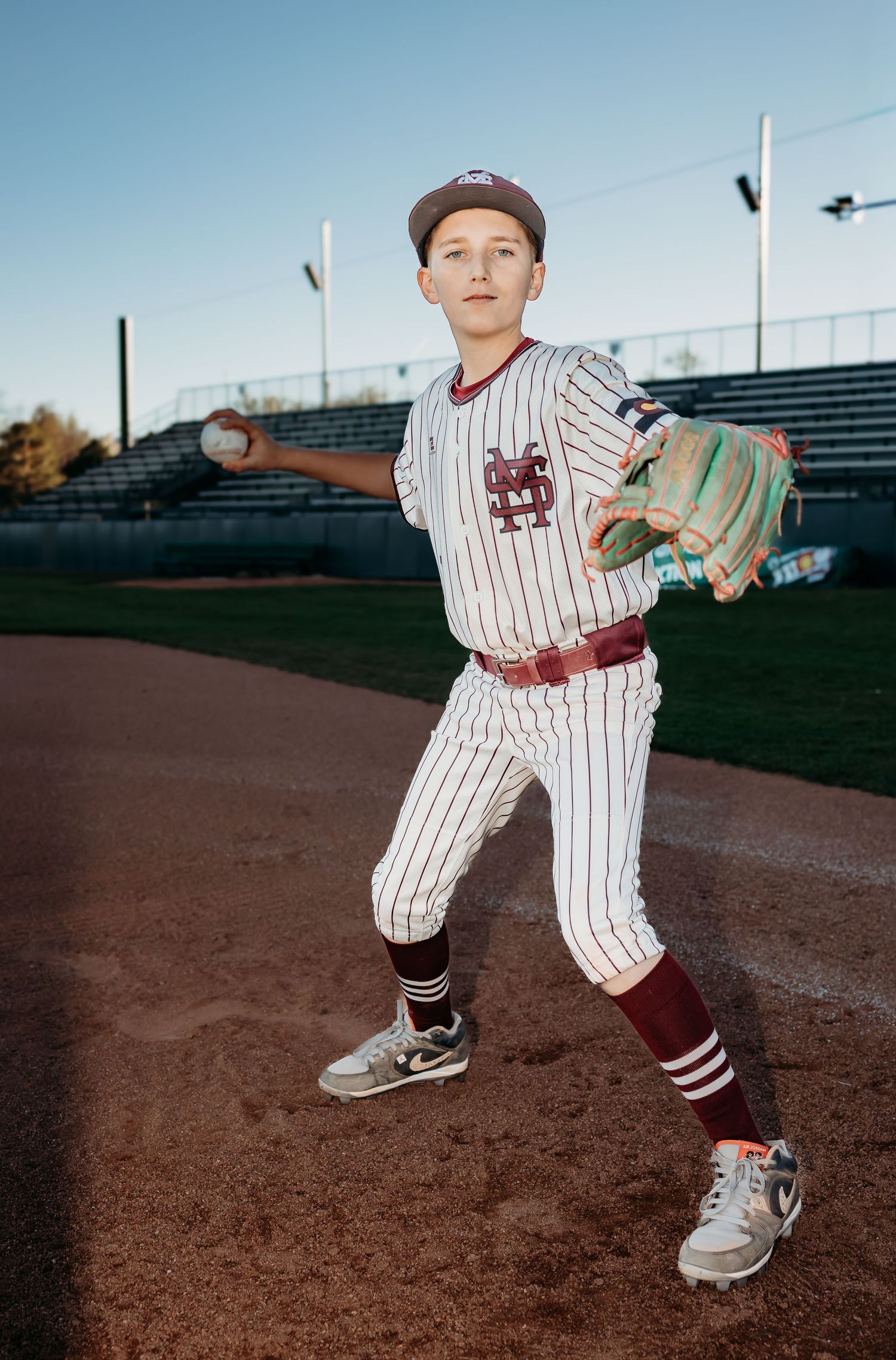 Young baseball player in a pinstriped jersey and maroon socks extends his glove while holding a ball, posing on a ballfield infield.