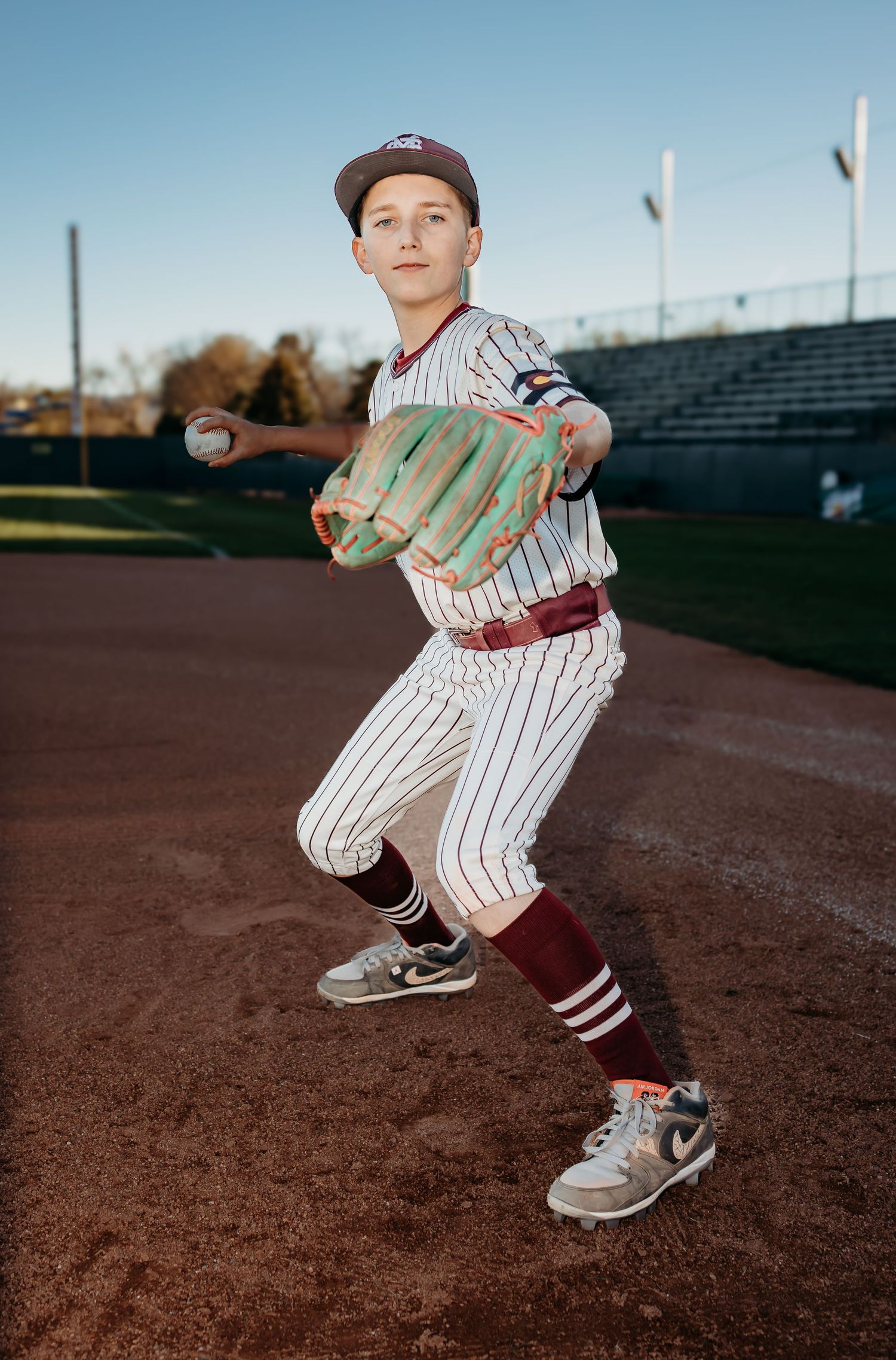 Boy in a pinstriped baseball uniform and maroon socks poses mid-throw with a green glove and ball on a sunlit infield.