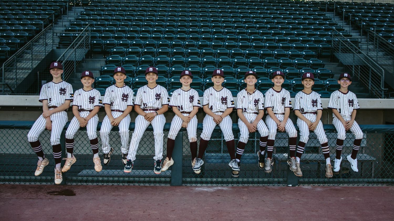 Team of young baseball players in matching white and maroon pinstriped uniforms sits in a row on a dugout fence in front of stadium seats.
