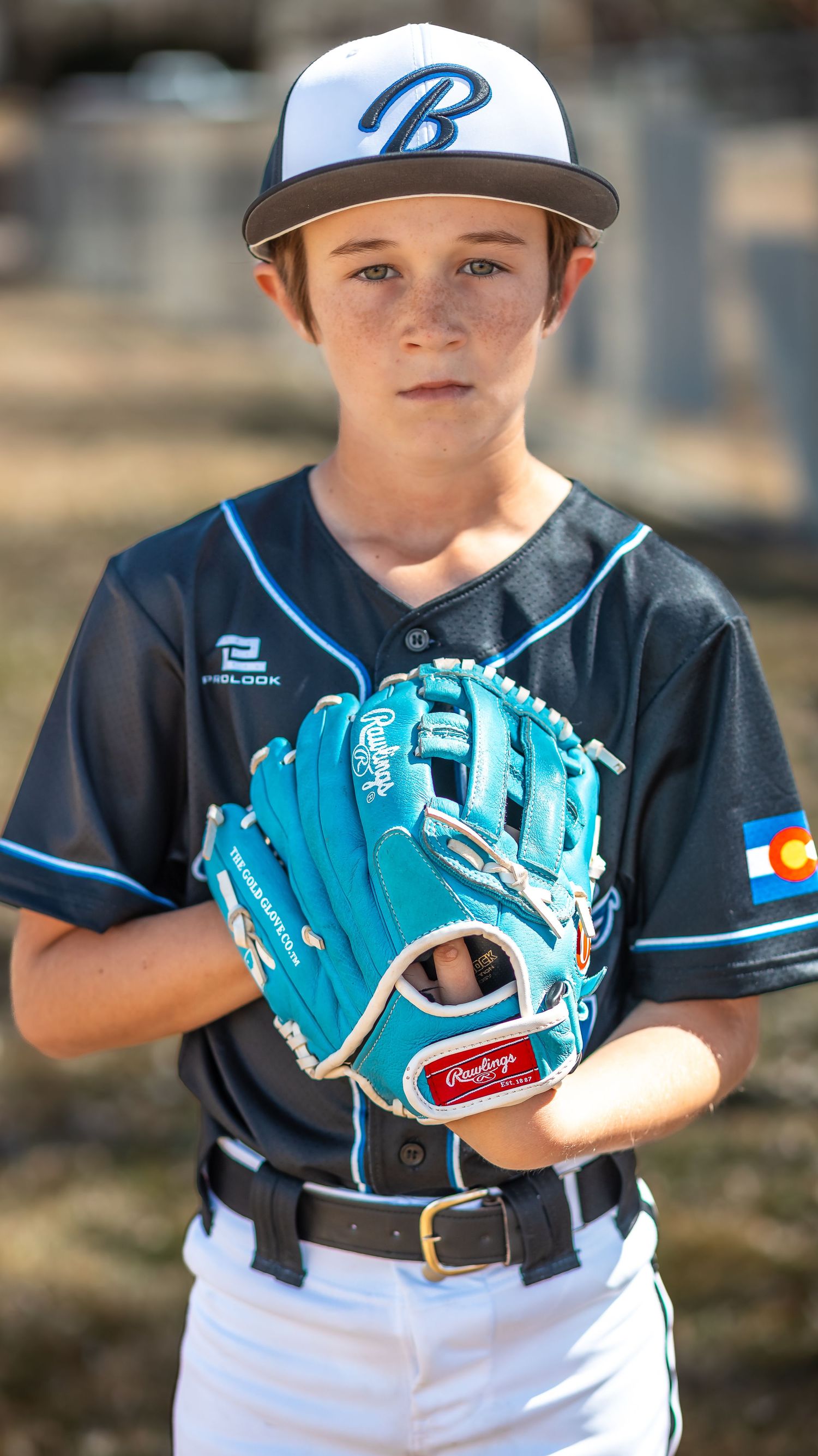 Young baseball player in a black jersey and white B cap holds a turquoise Rawlings glove while posing outdoors.