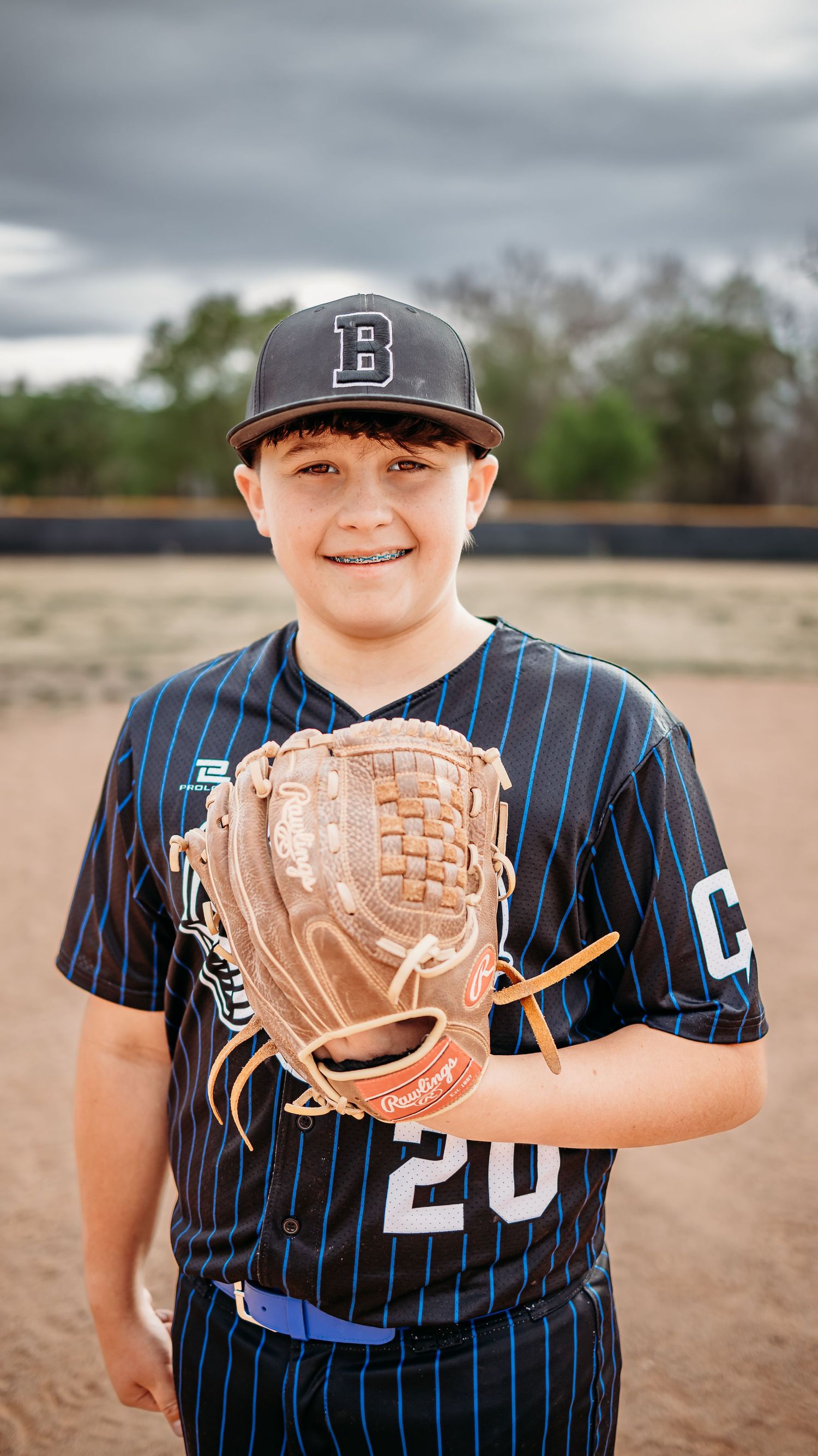 A smiling young baseball player in a black pinstripe jersey and cap holds a Rawlings glove on a dirt infield under cloudy skies.