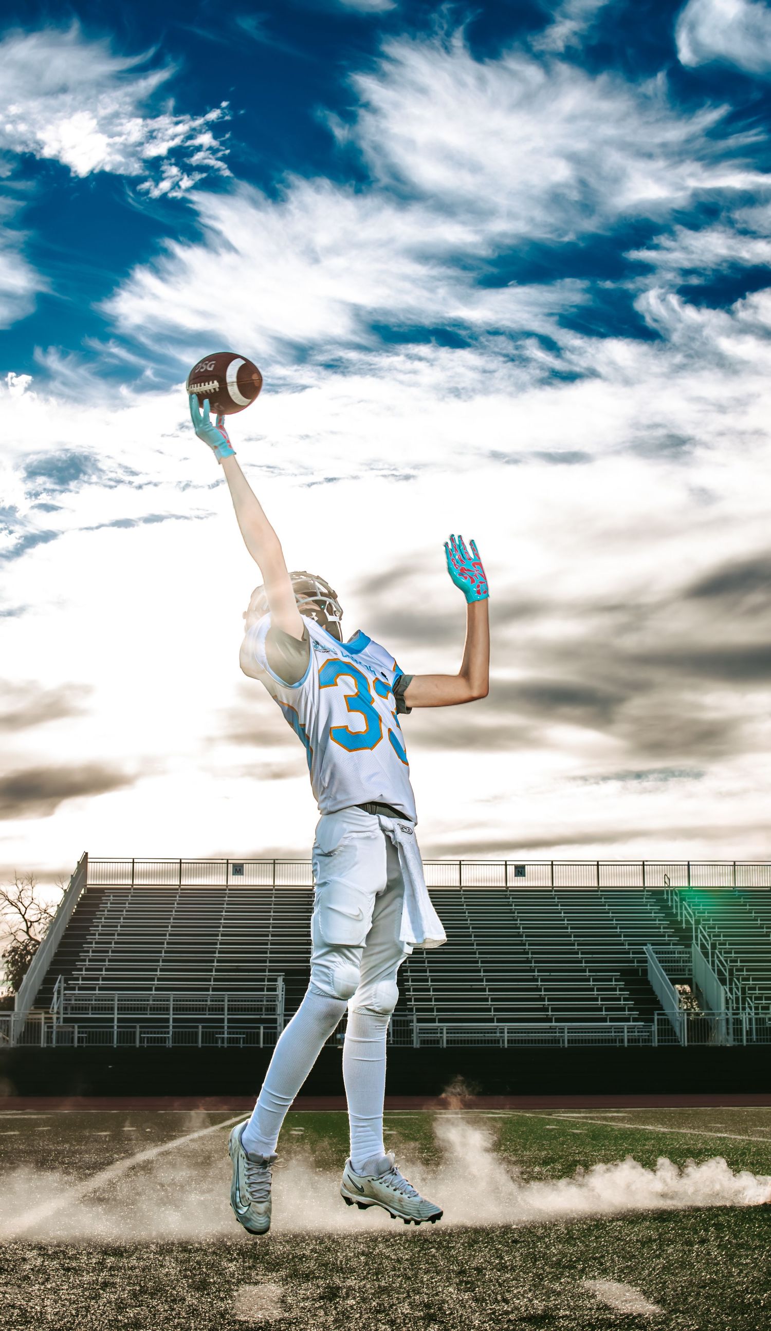 A youth football player in a white and teal uniform leaps with one arm stretched to catch a football against a bright cloudy sky.
