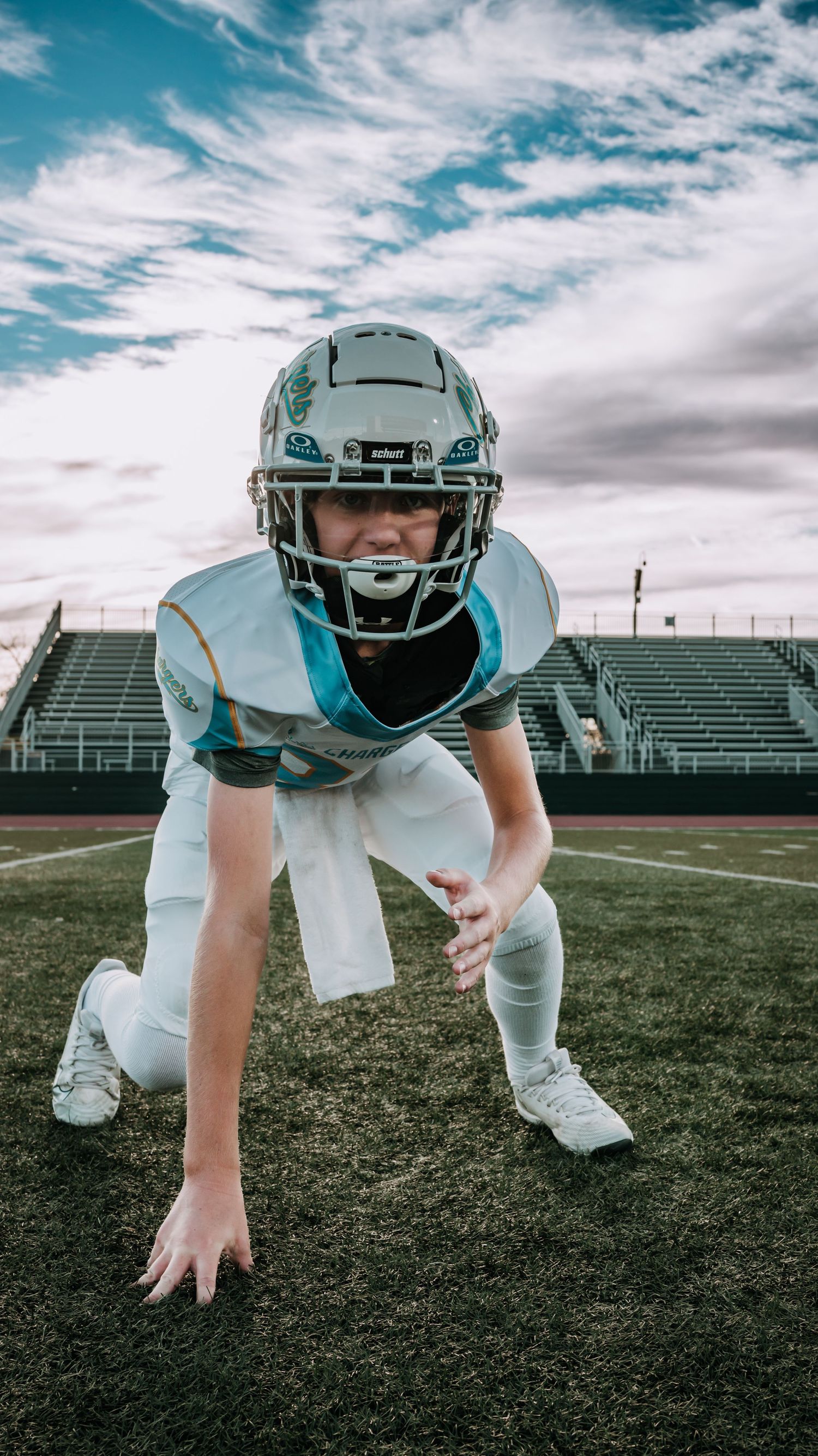 A youth football player in a white helmet and teal-trimmed jersey crouches in a three-point stance on a turf field before empty bleachers.