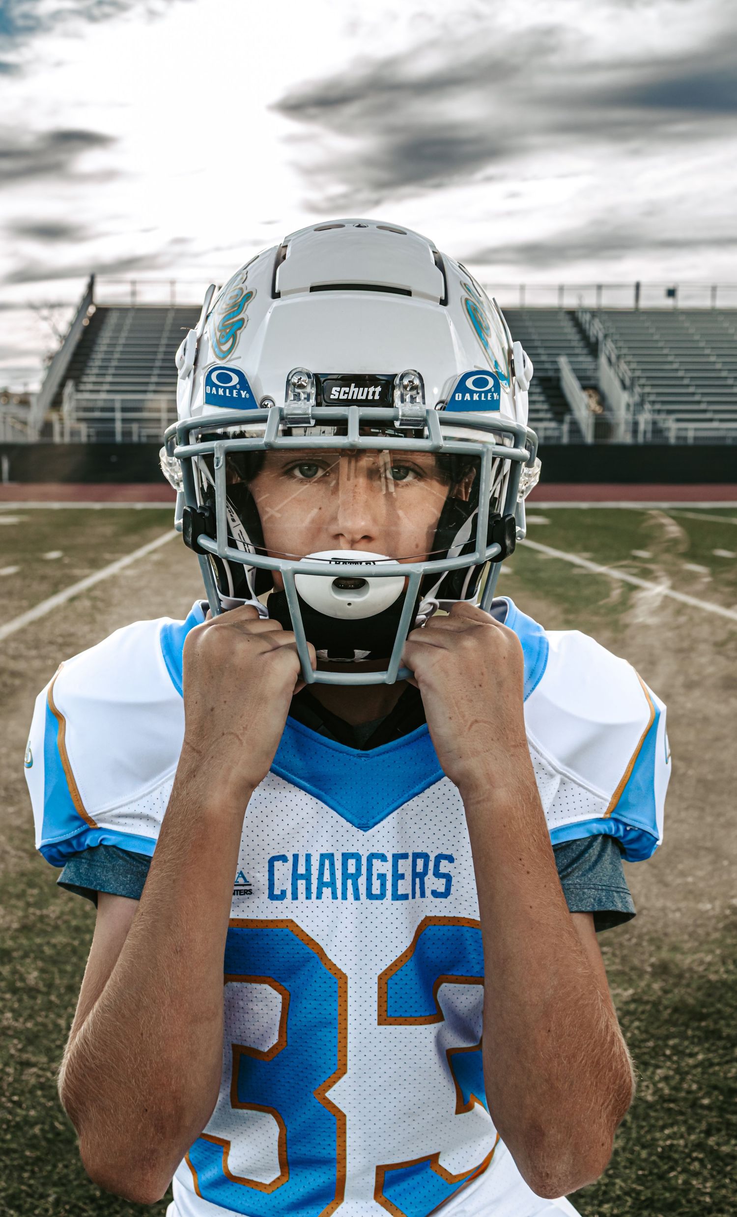 Youth football player in a white and blue Chargers jersey and helmet gripping his chin strap on a stadium field under cloudy skies.