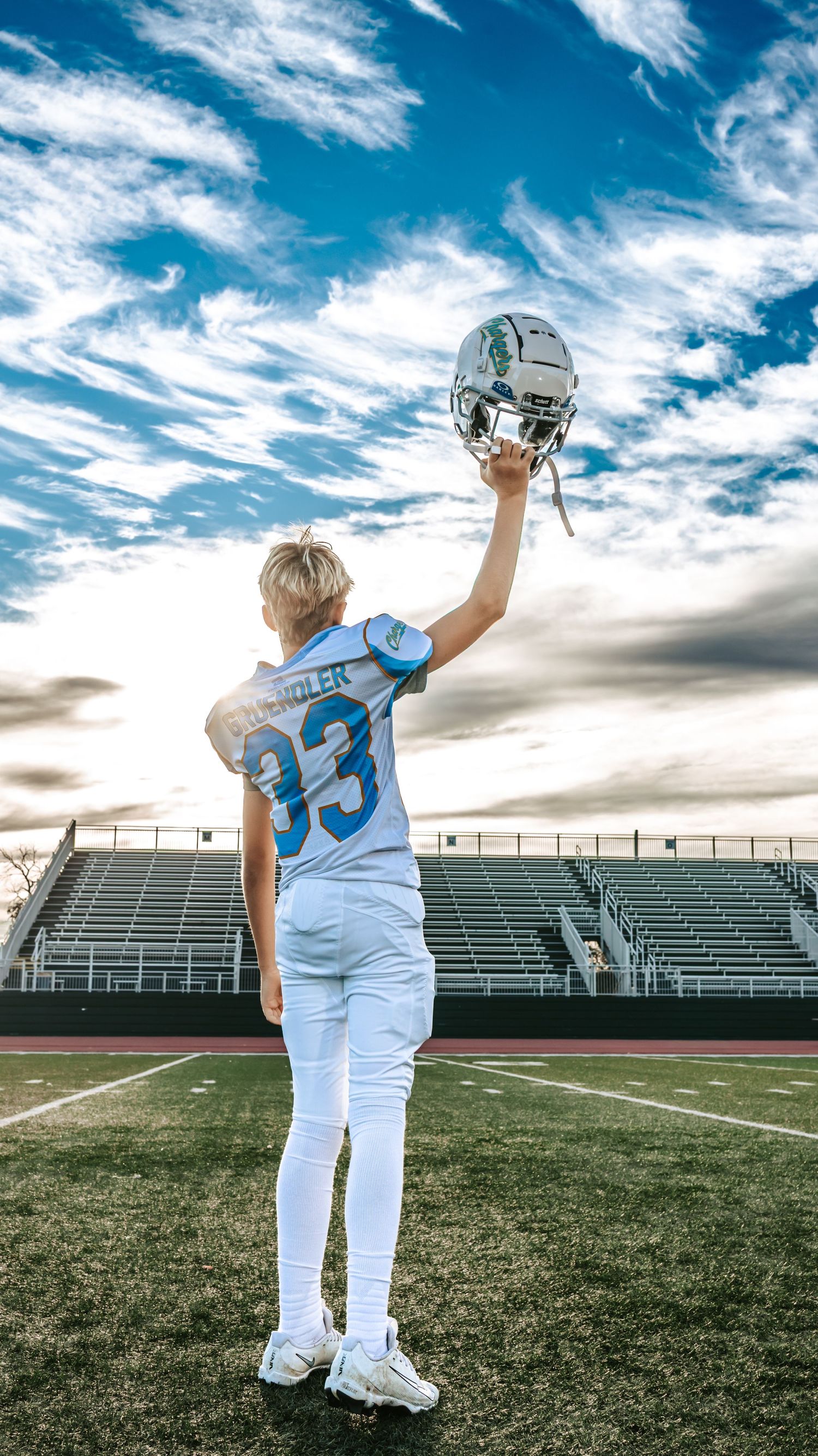 Chargers football player in a number 33 jersey raises his helmet toward a bright blue sky with wispy clouds above stadium bleachers.