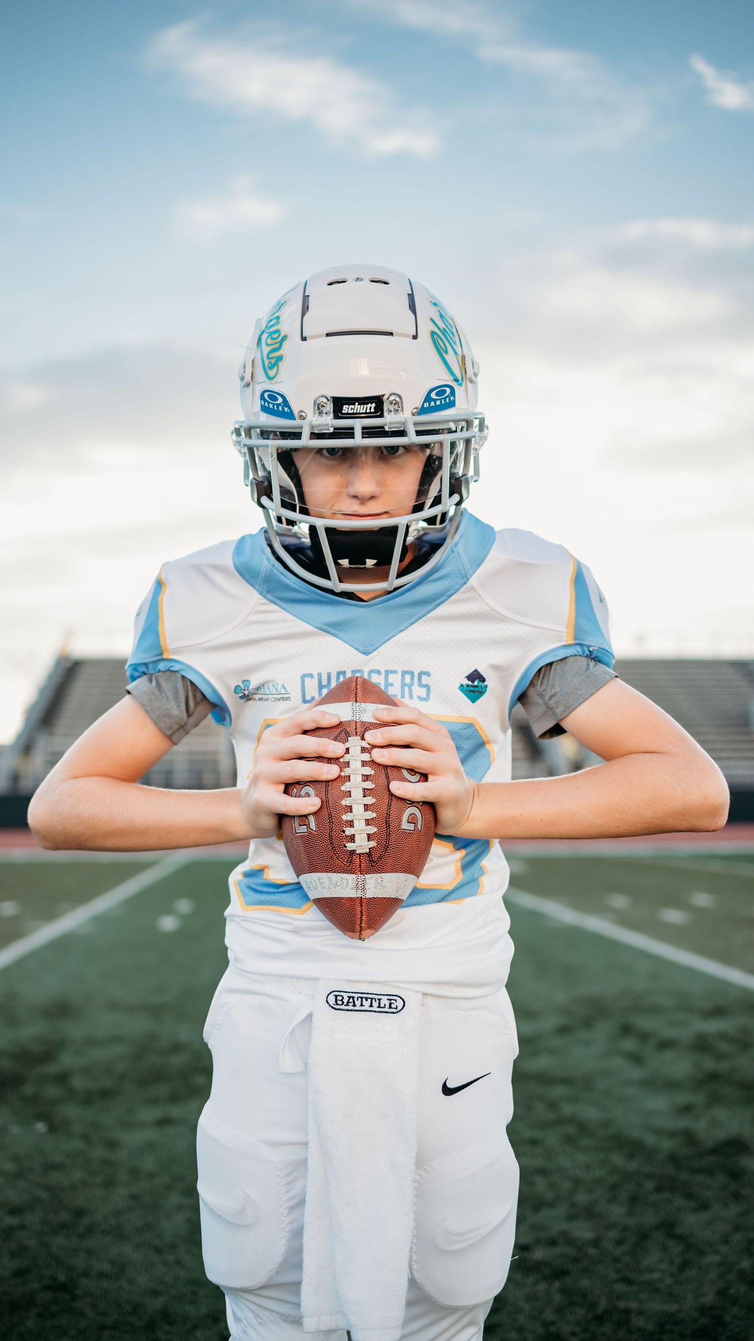 Young Chargers football player in a white helmet and jersey holds a football at his chest while standing on a green turf field.
