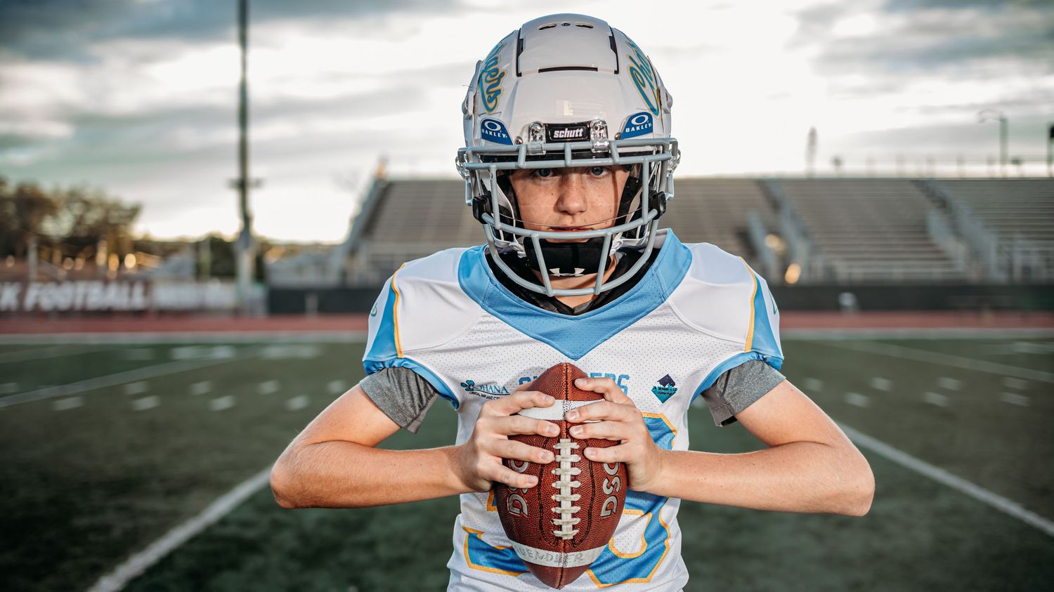 Youth football player wearing a white and light blue Chargers uniform and helmet holds a football with both hands on the field.