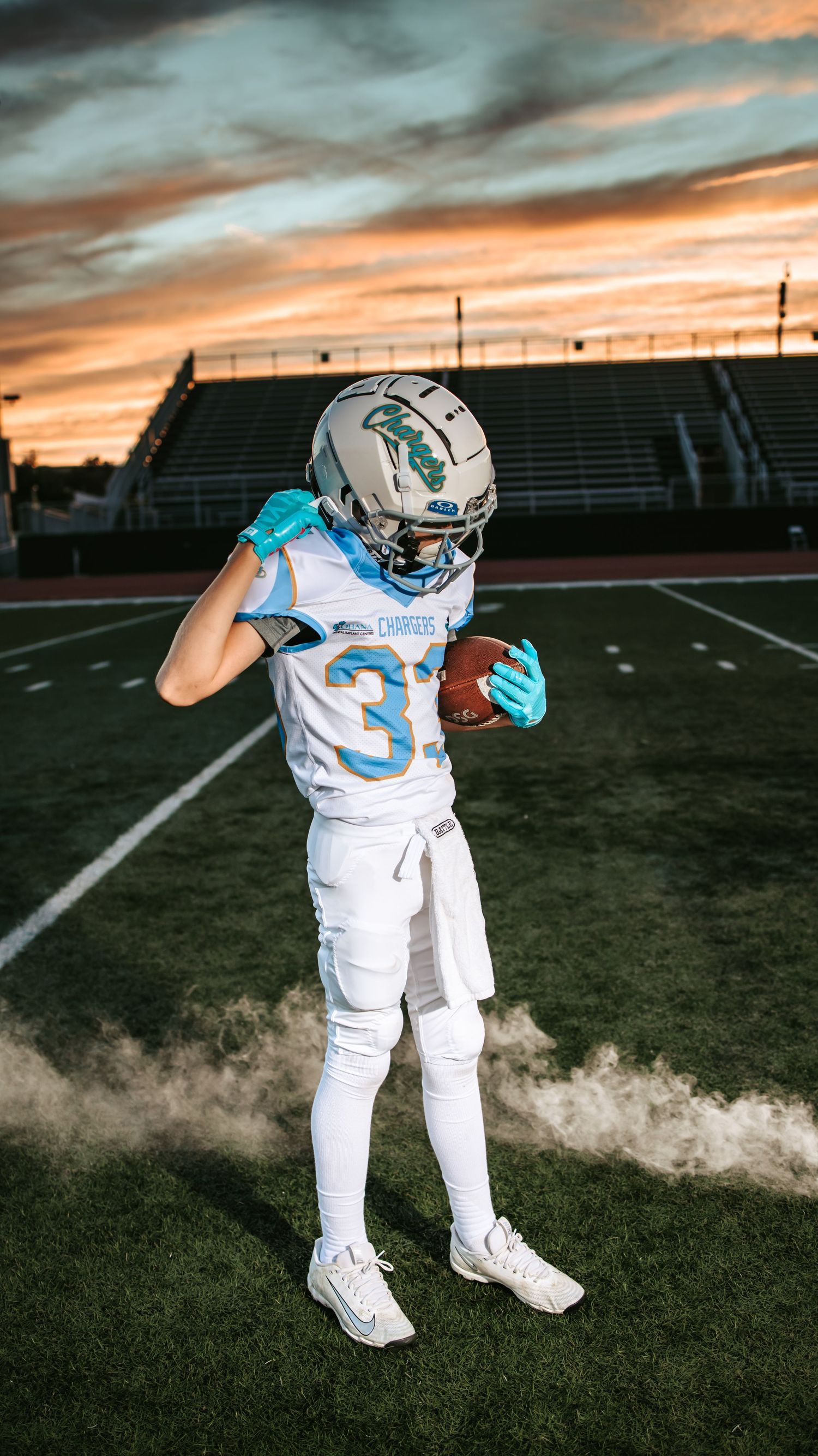 Young Chargers football player in teal gloves looks down at the football in his hand on a field beneath a colorful sunset sky.