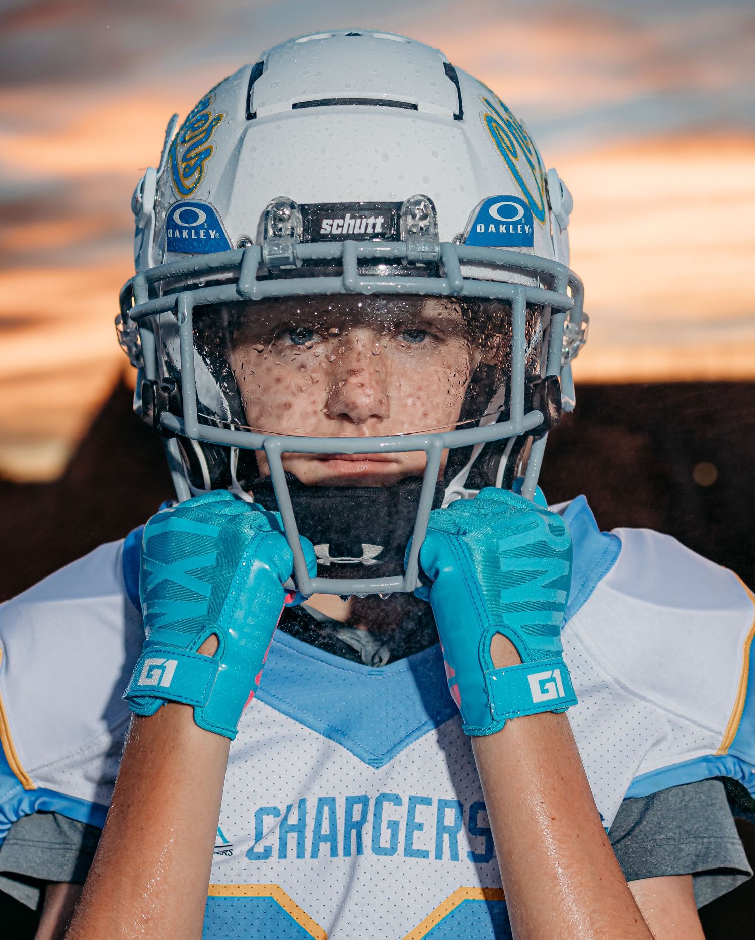 Close-up of a youth football player in a white Chargers helmet and teal gloves with sweat-dampened face and a golden sunset behind him.