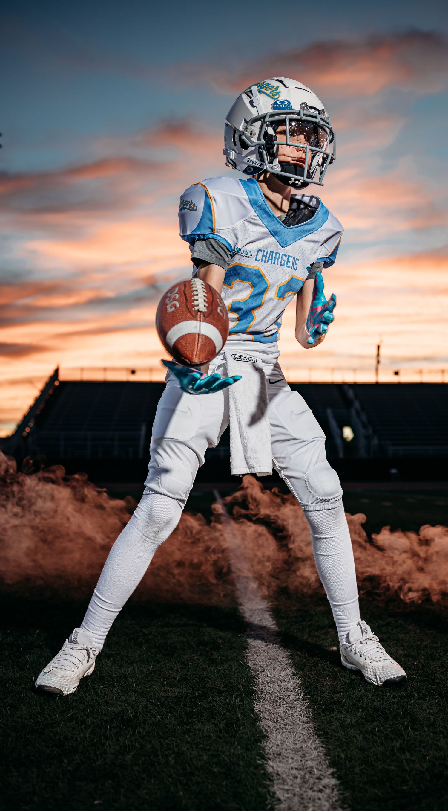 Chargers youth football player in teal gloves tosses a football while standing in a wide stance with dust swirling under a vivid sunset.