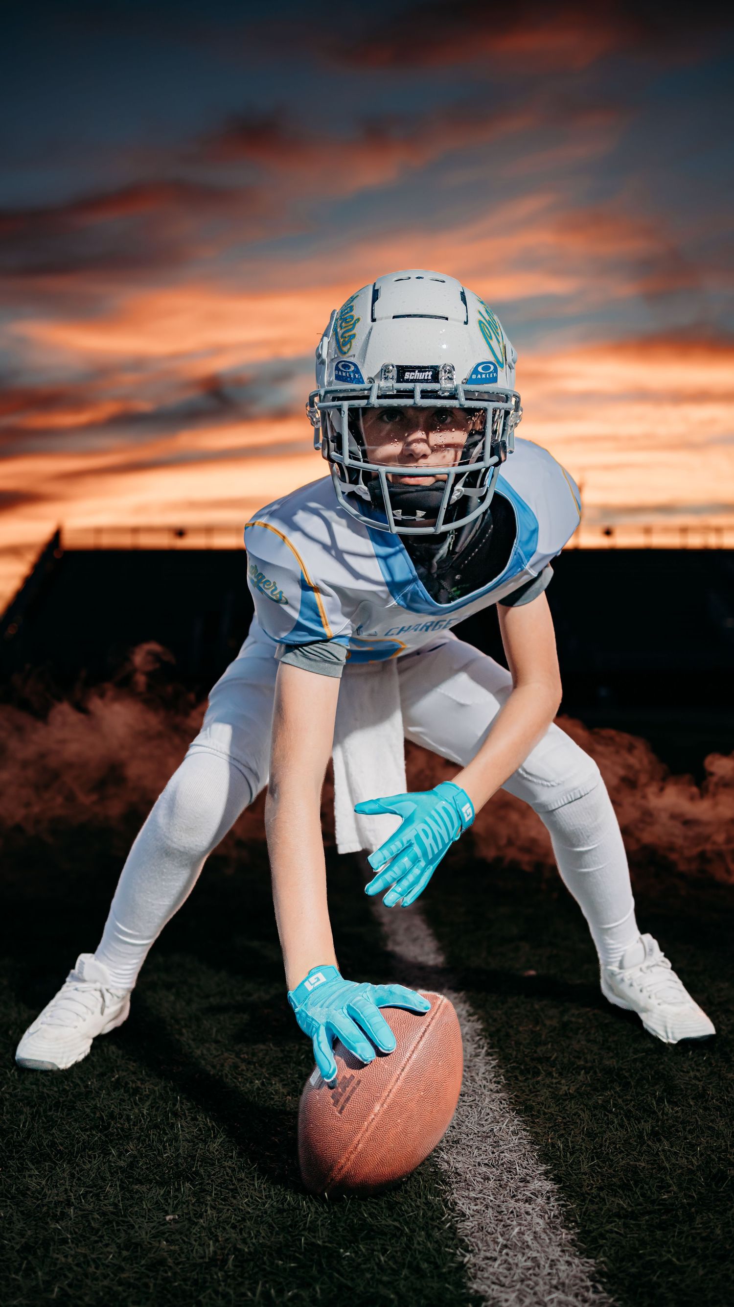 Young Chargers football player in a helmet and teal gloves crouches in a ready stance with a football on the turf at sunset.