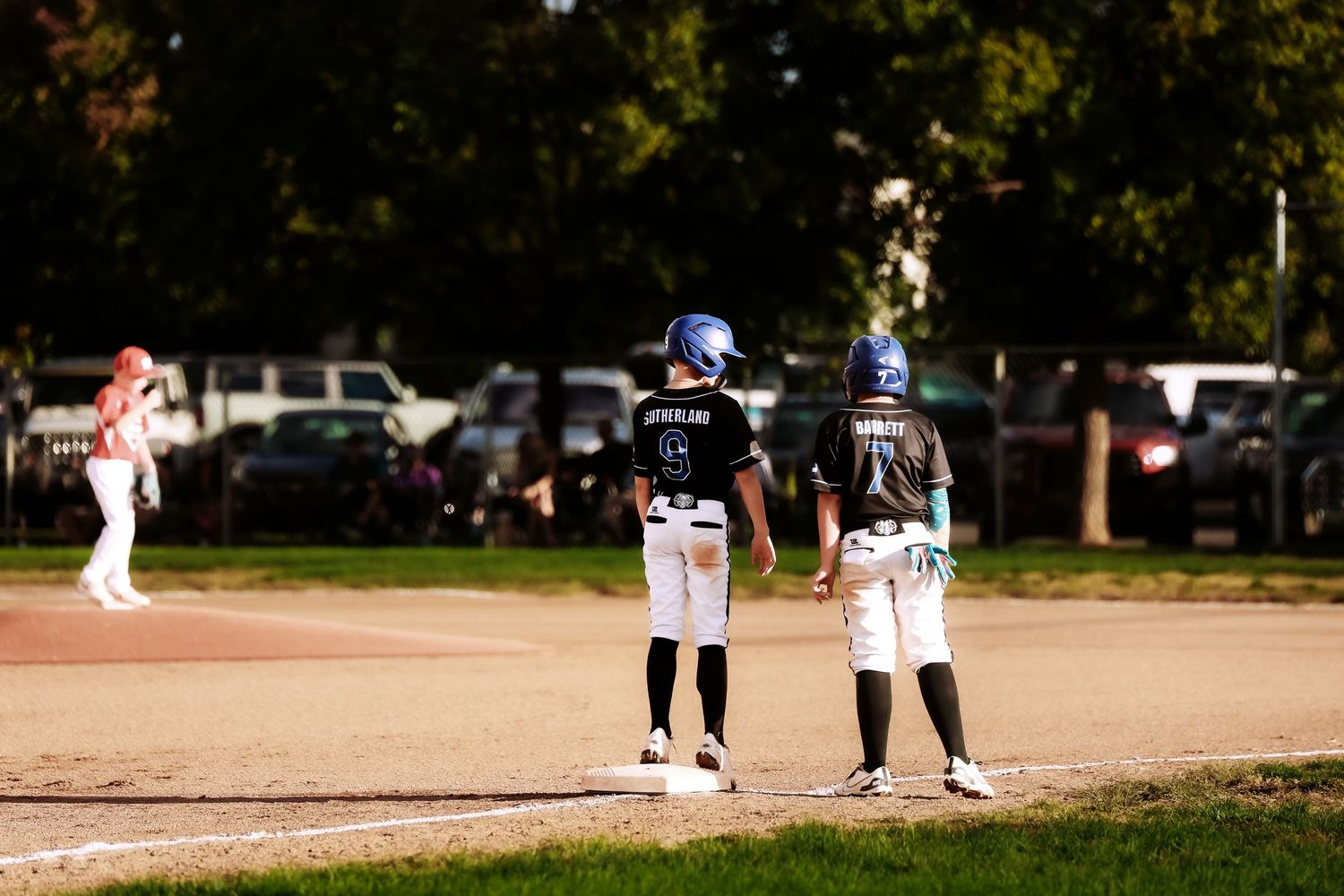 Two youth baseball players in helmets stand near a base during a game while a runner in a pink helmet watches from the background.