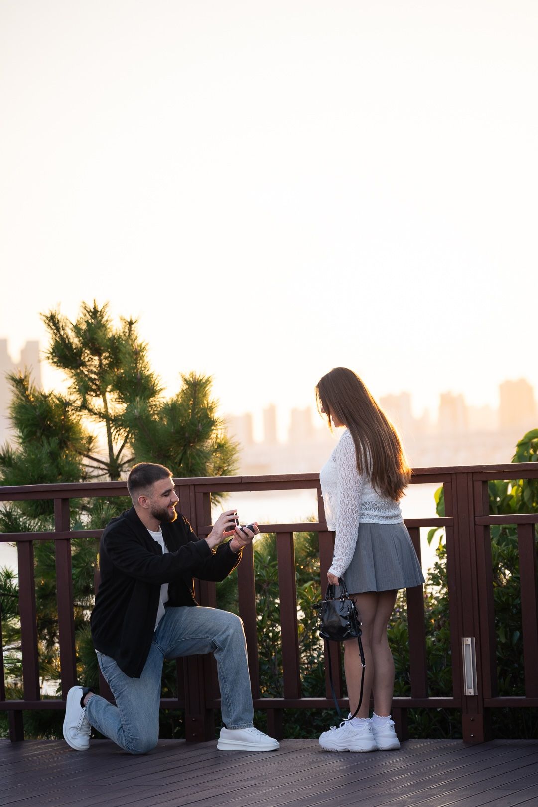 Man proposes on one knee to woman on wooden deck at sunset with city skyline in background.