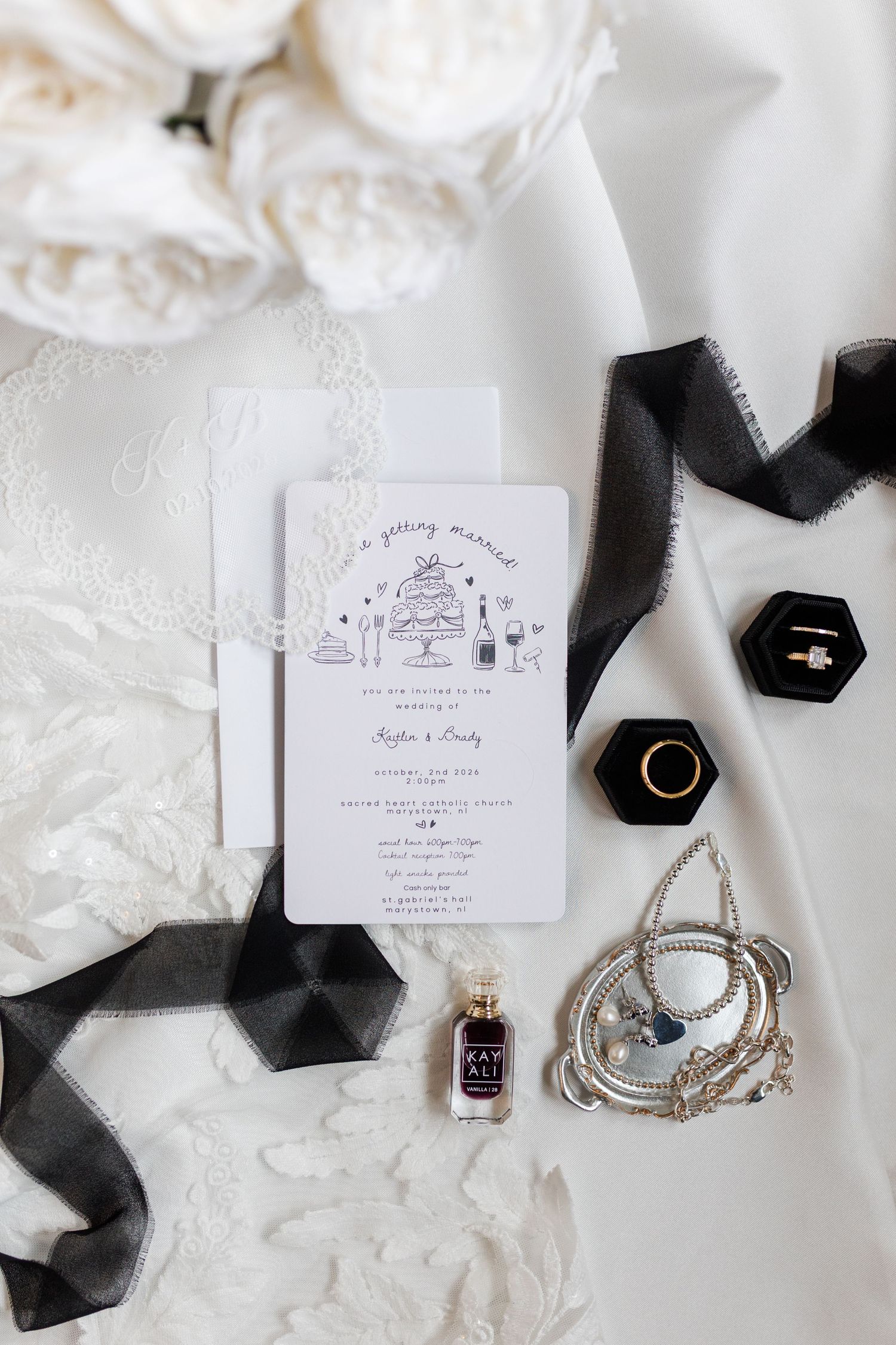 Wedding invitation with cake illustration beside black ribbons, two ring boxes, a perfume bottle, jewelry, white roses and lace.