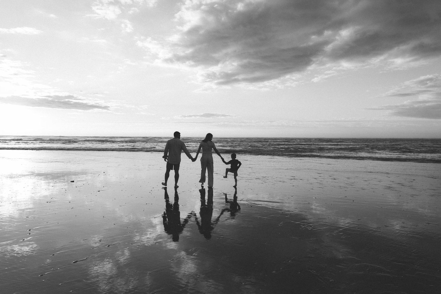 Silhouette of two adults and a child holding hands on a wet beach at sunset, with reflections on the sand and waves in black and white.