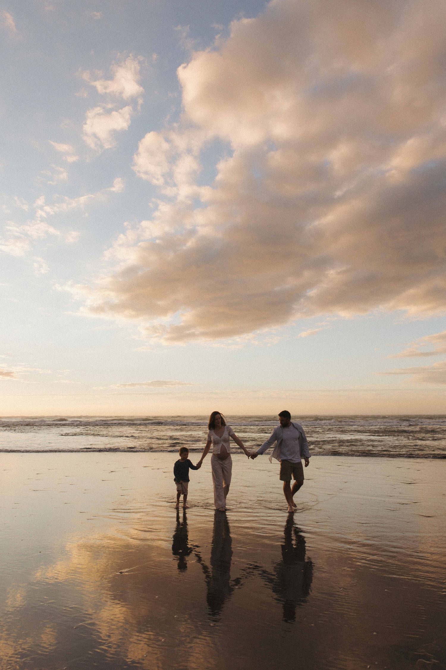 Family of three holding hands and walking on a wet sandy beach at sunset, with golden clouds reflected in the shimmering shoreline.