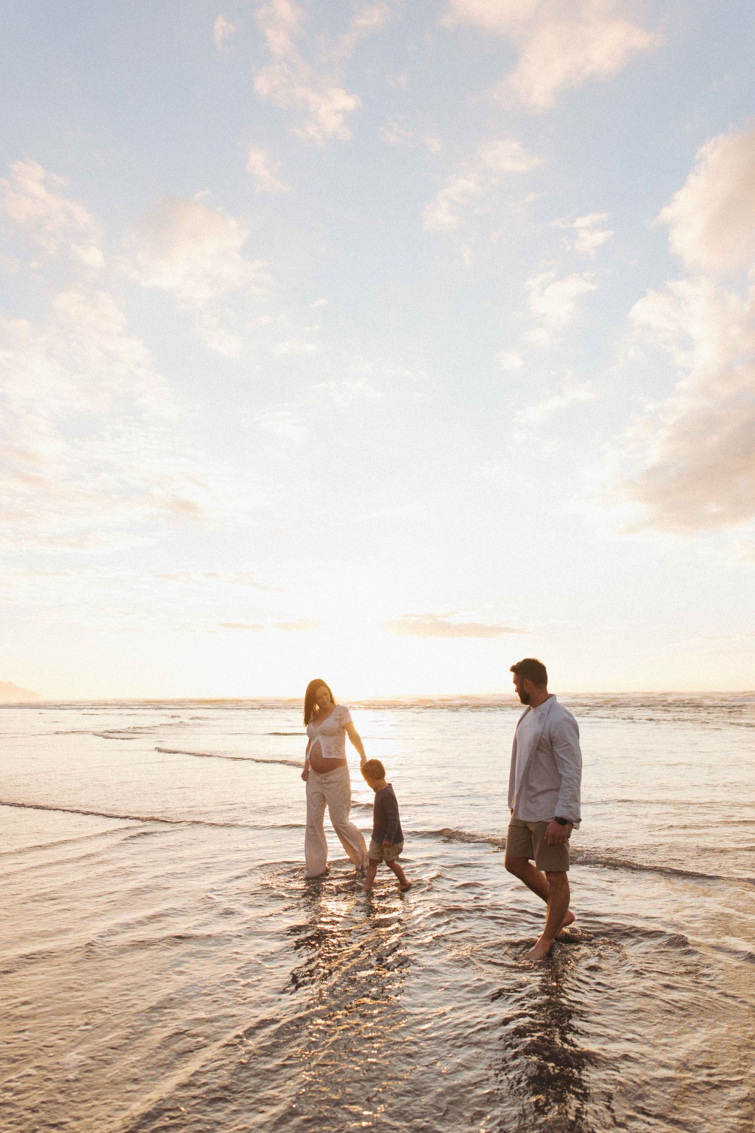 A family of three walks barefoot along the shallow waves of a beach at sunset, with soft golden light and clouds filling the sky.