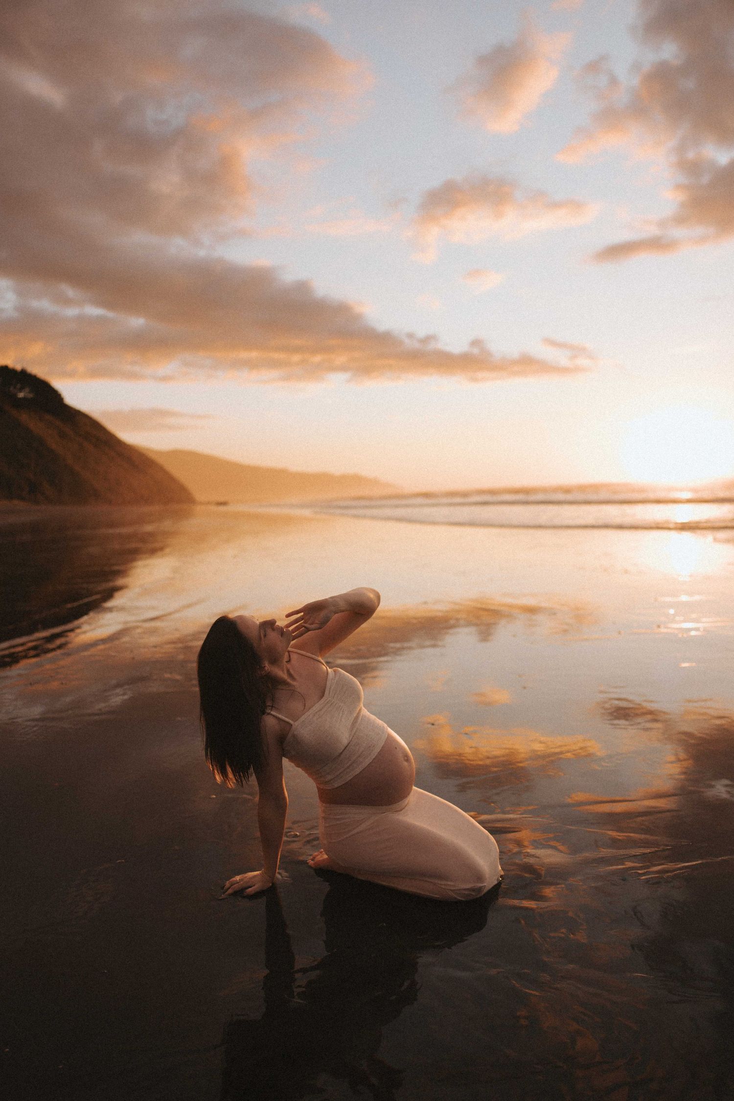 Pregnant woman in a flowing cream outfit kneels on wet sand at sunset, tilting her head back as golden clouds reflect on the shoreline.