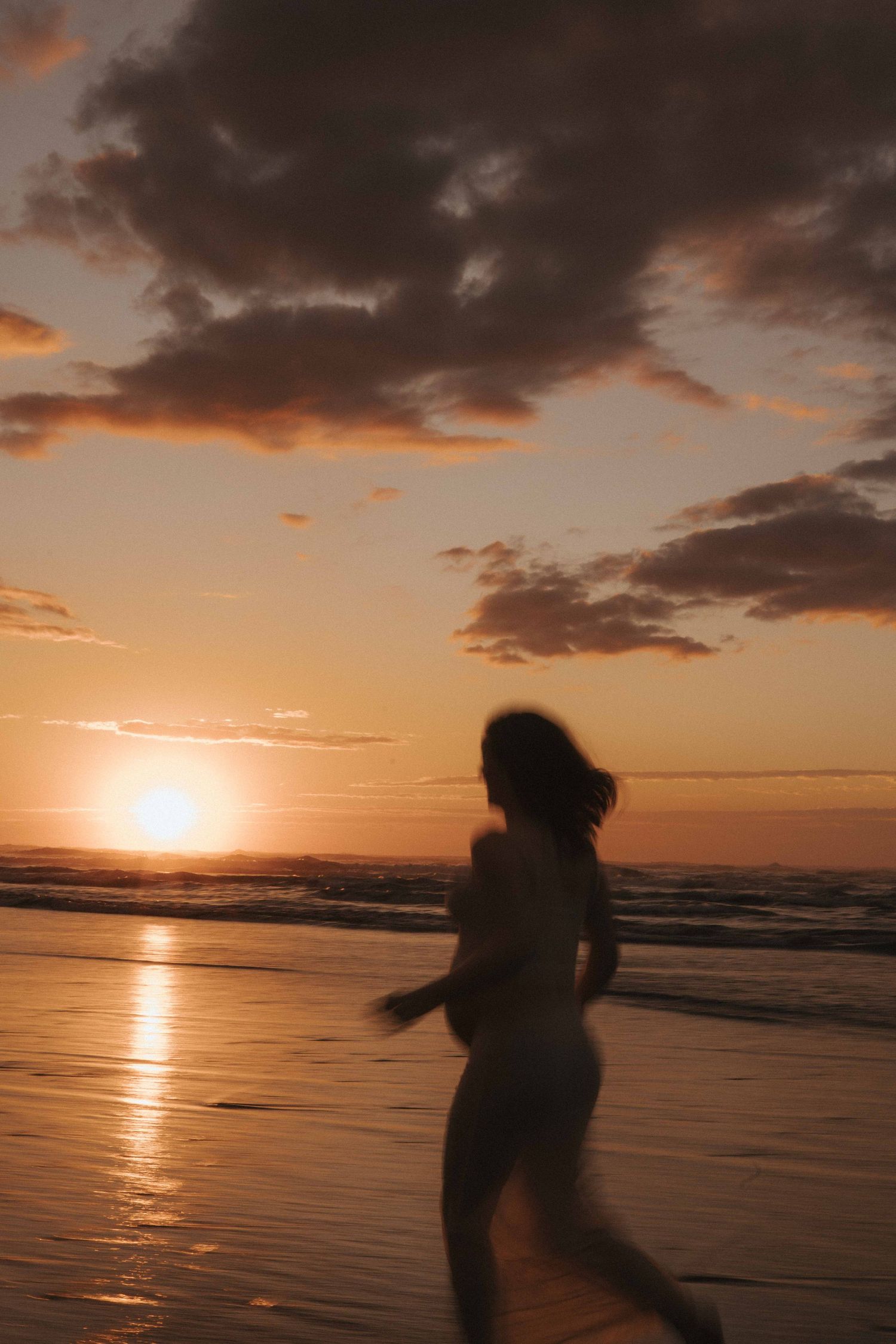Silhouette of a woman running along a wet sandy beach at sunset with golden sun reflections on the water and dramatic clouds overhead.