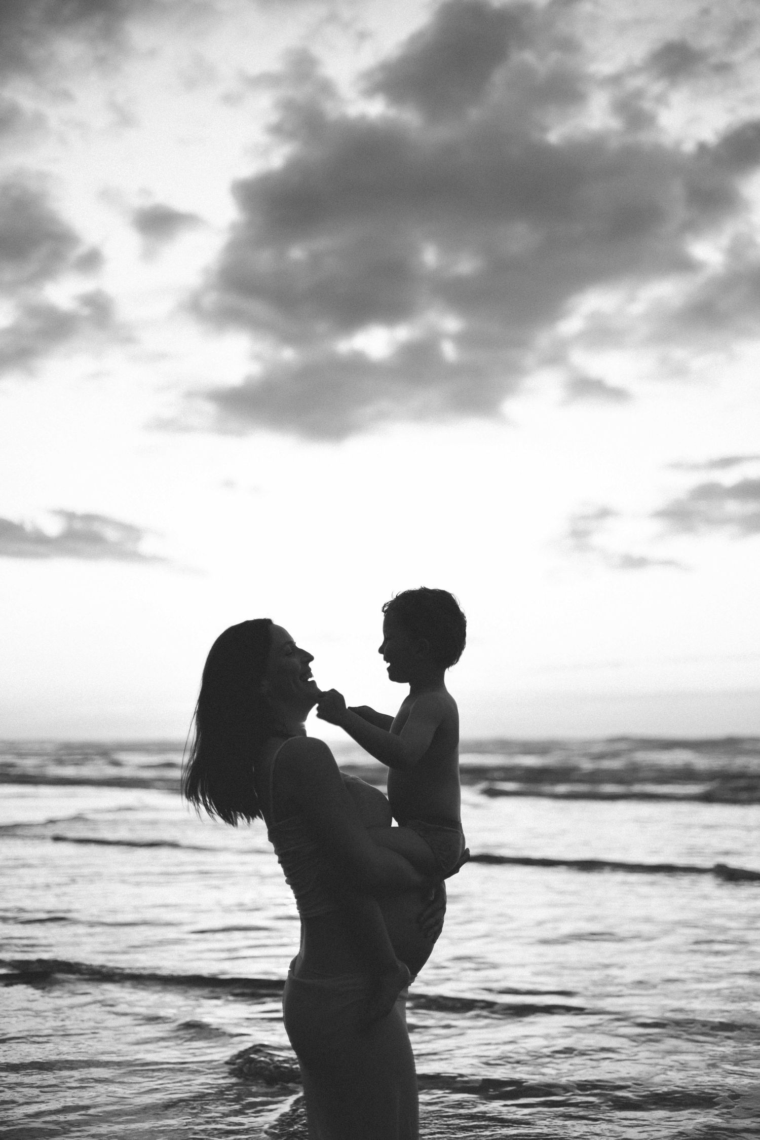Black and white silhouette of a mother lifting her smiling toddler at the ocean shore under a cloudy sky at sunset.