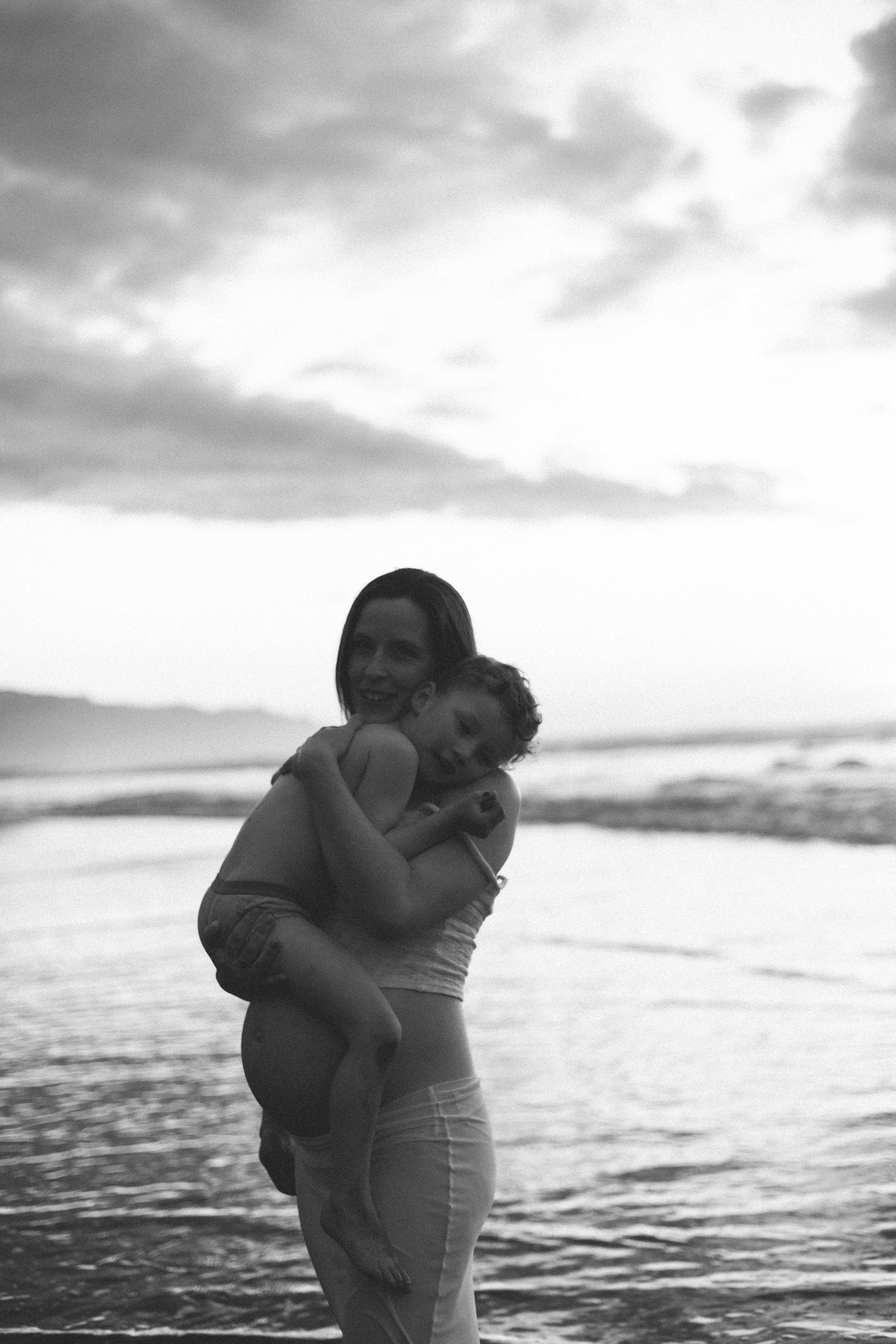Black and white photo of a smiling woman holding a child in her arms while standing in shallow ocean water at sunset.