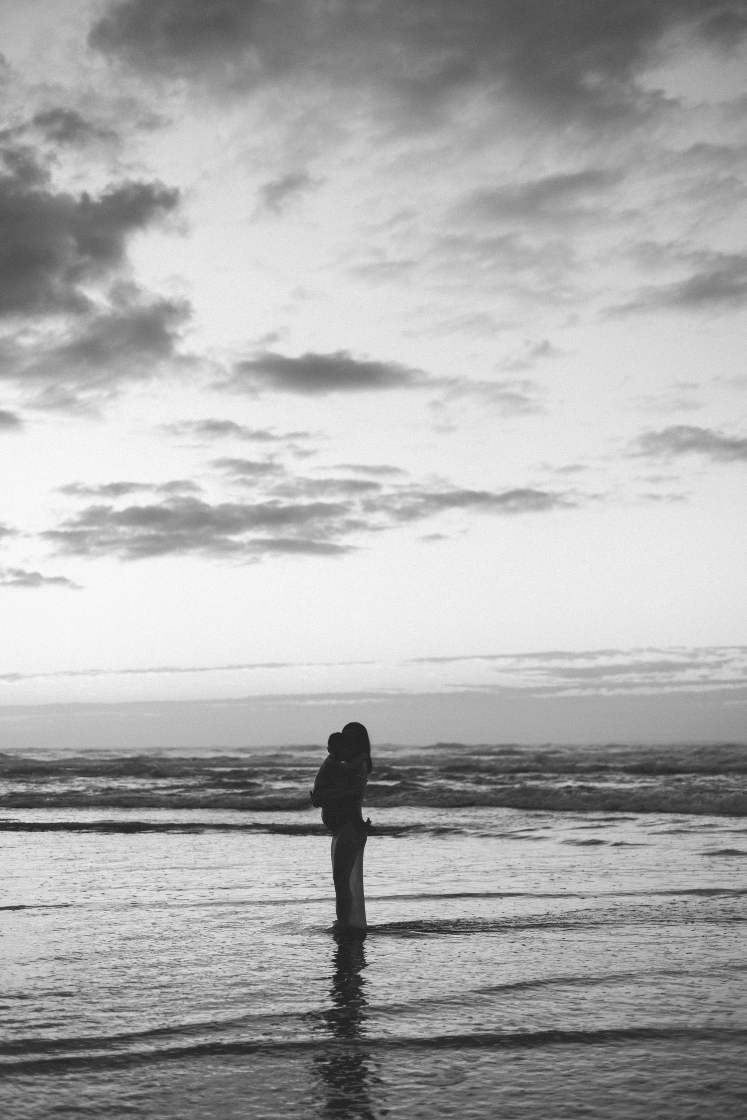 Black and white photo of a person standing in shallow ocean water at dusk, holding a child under a dramatic cloudy sky.