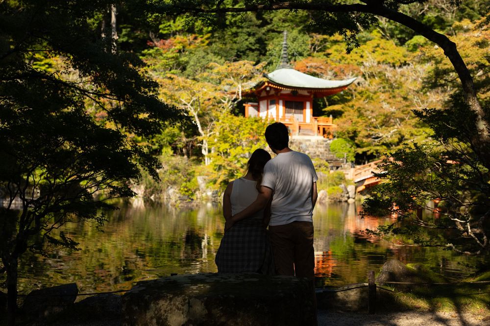 Couple standing together by a tranquil pond, gazing at a traditional Japanese temple pavilion surrounded by lush green and autumn foliage.