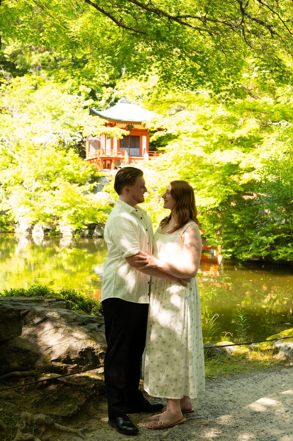 Couple embracing in dappled sunlight beside a tranquil pond with a red Japanese pavilion framed by lush green maple trees.