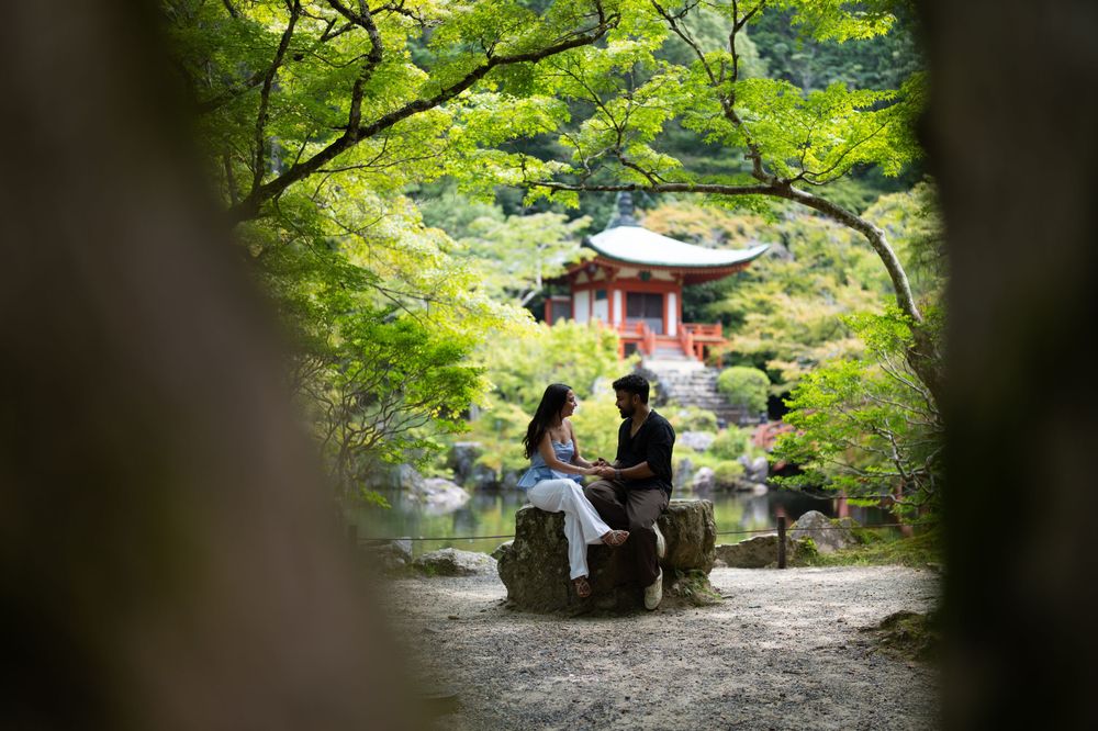 Couple sitting together on a stone seat beside a pond, framed by green maple leaves, with a red Japanese temple pavilion in the background.