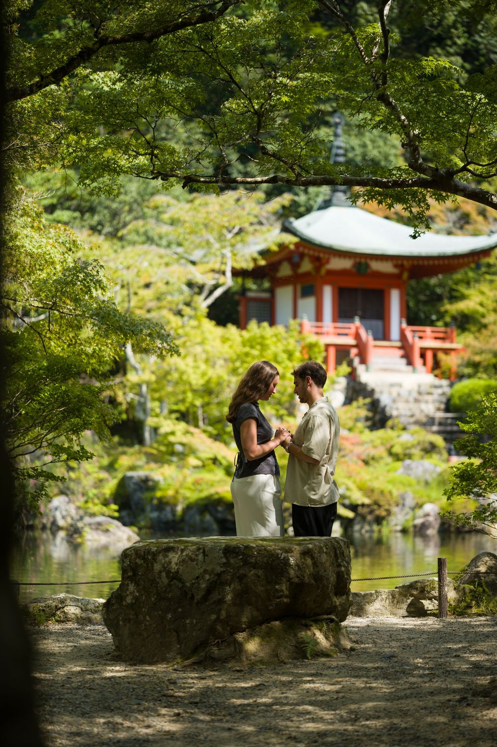 Couple holding hands beneath lush green trees beside a tranquil pond, with a red and white Japanese pagoda-style pavilion in the background.