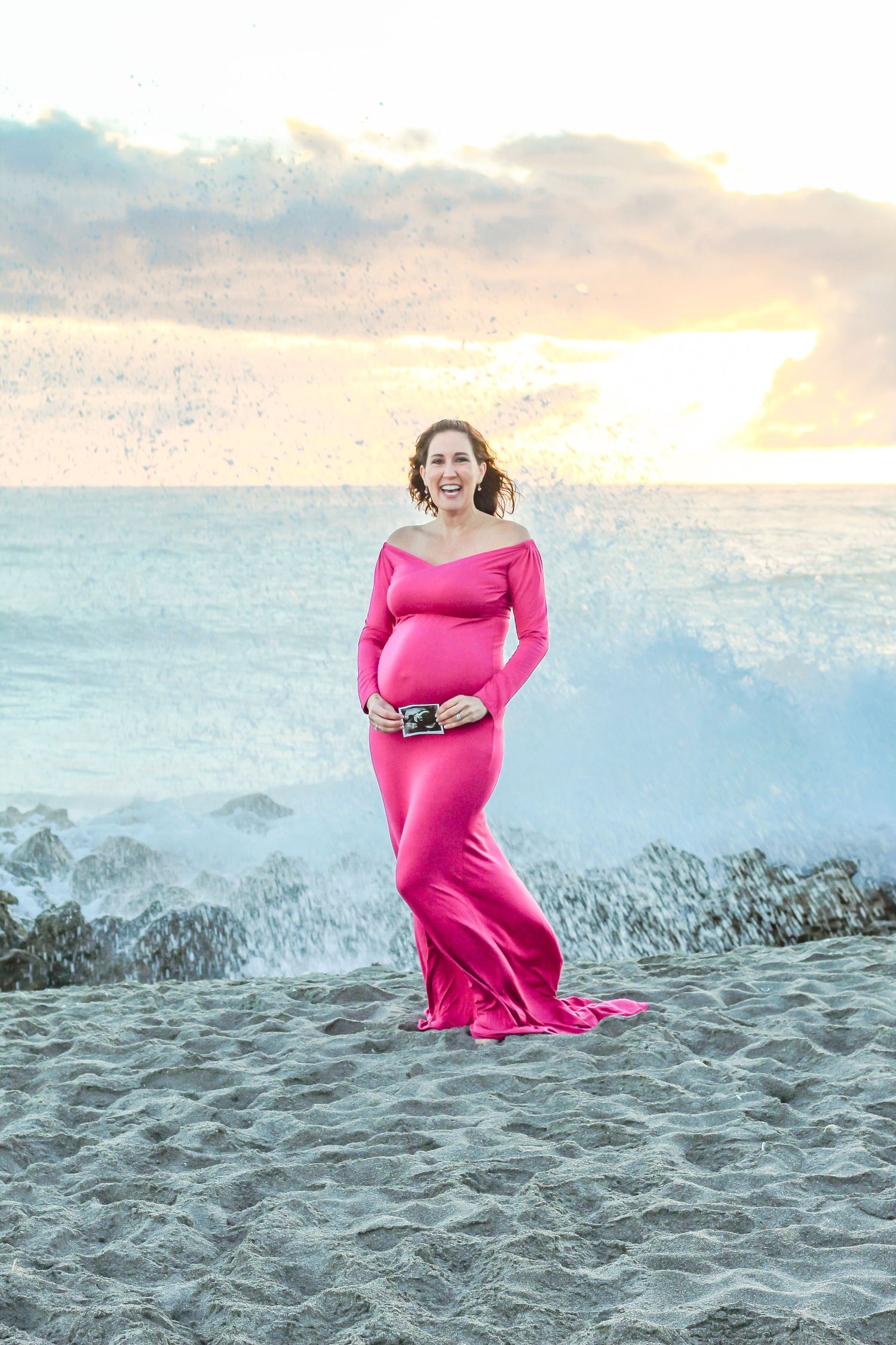 Smiling pregnant woman in pink off-shoulder gown poses on sandy beach at sunset with ocean waves crashing behind her at Blowing Rocks, FL