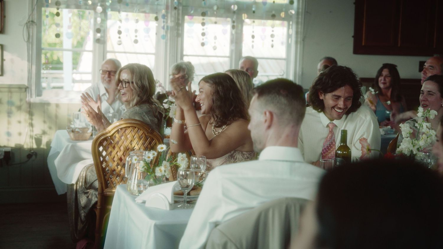 Guests celebrate at a wedding reception, clapping and laughing around a flower-decorated table indoors.