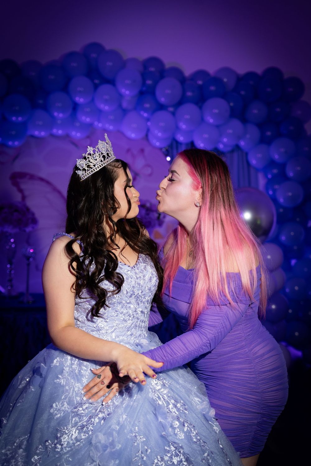 Two girls pose at a quinceañera, one in a blue ball gown with tiara, the other in purple, with balloon backdrop.