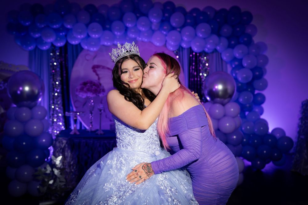 Two women celebrate a quinceañera, one in a blue ball gown with crown receives a kiss at a purple balloon decorated party.