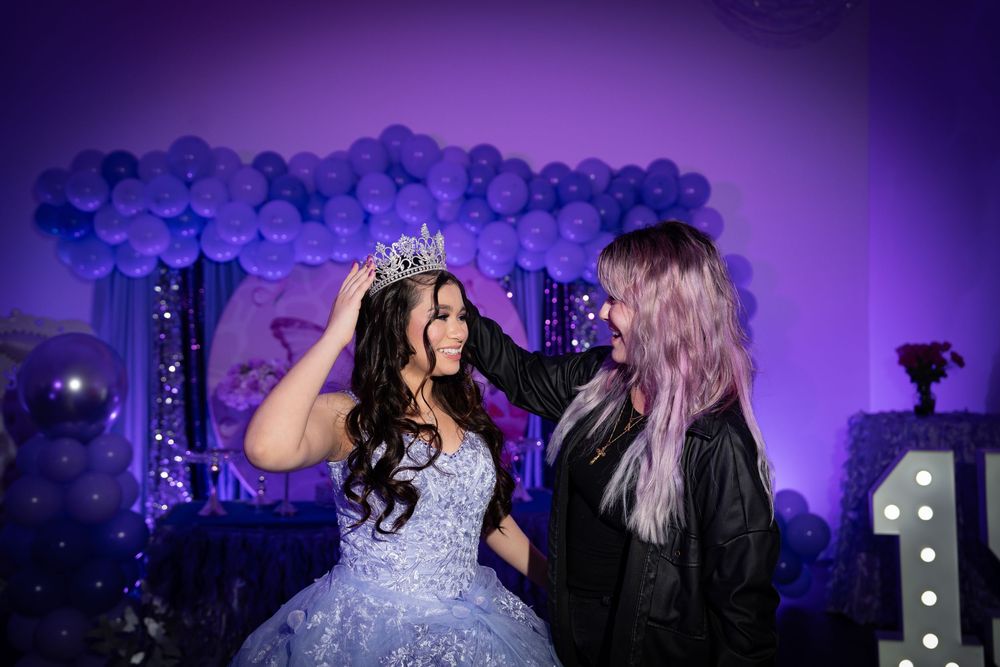 A young woman in a blue quinceañera gown receives a tiara crown at a purple balloon-decorated celebration.