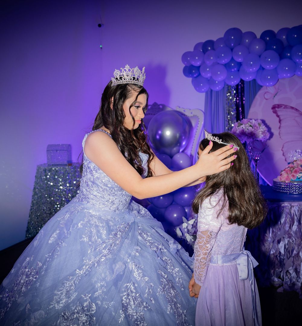 A quinceañera in a blue gown places a tiara on a young girl at a purple balloon-decorated celebration.