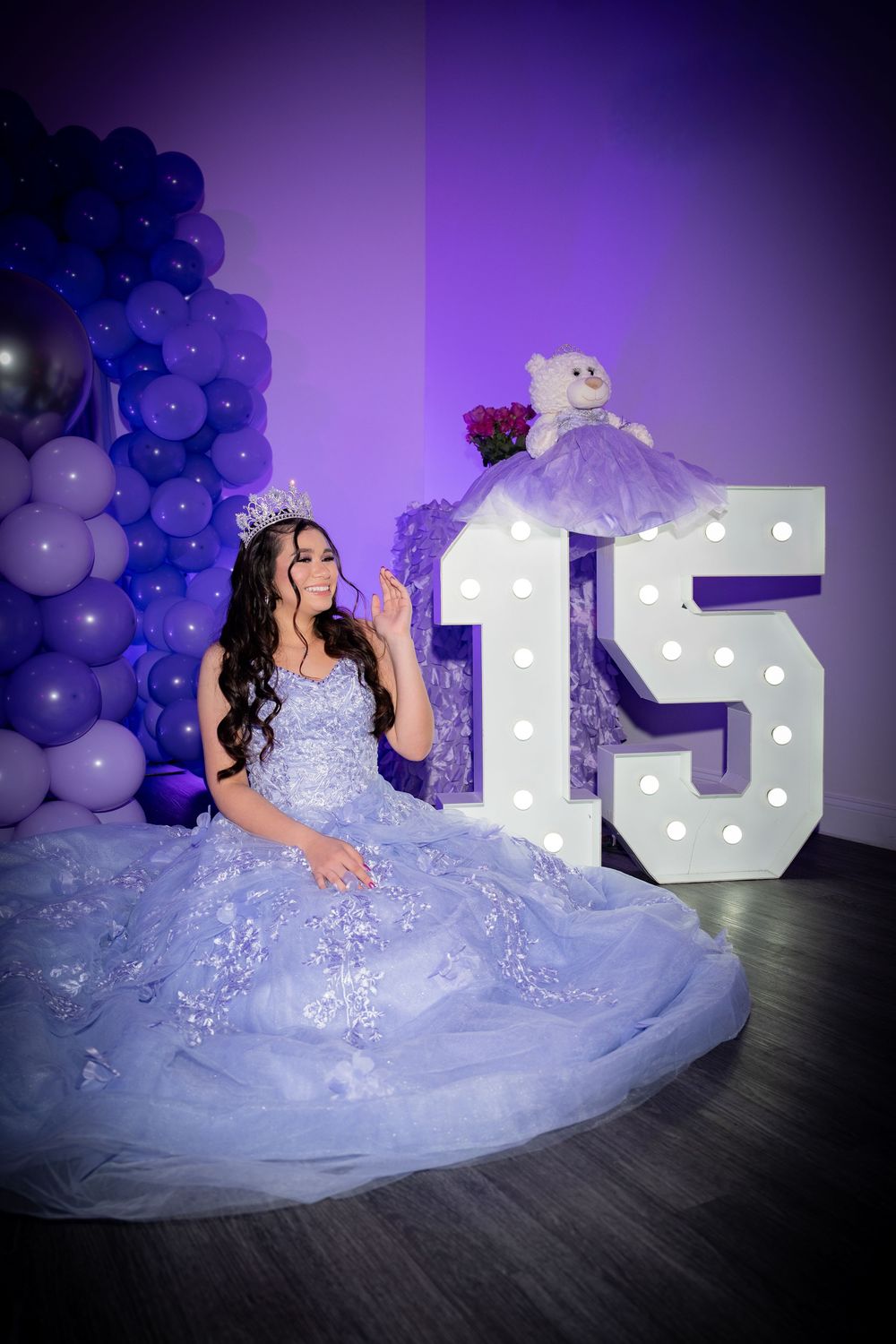 A young woman in a lavender ball gown and crown sits beside illuminated number 15 at a quinceañera celebration.