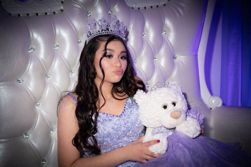 Young woman in lavender quinceañera gown and crystal crown holds white teddy bear on tufted white throne.