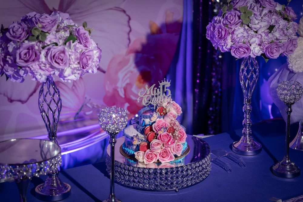 Elegant quinceañera cake with pink roses on a crystal stand, flanked by lavender floral arrangements on a blue-lit table.