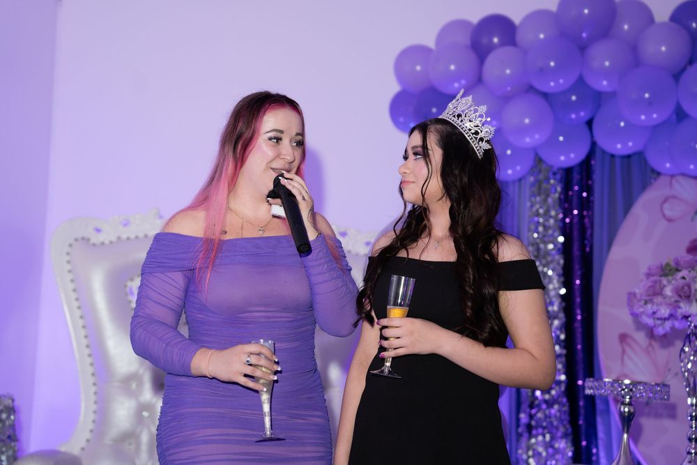 Two women celebrate at a purple-themed quinceañera party, one wearing a crown and holding champagne.
