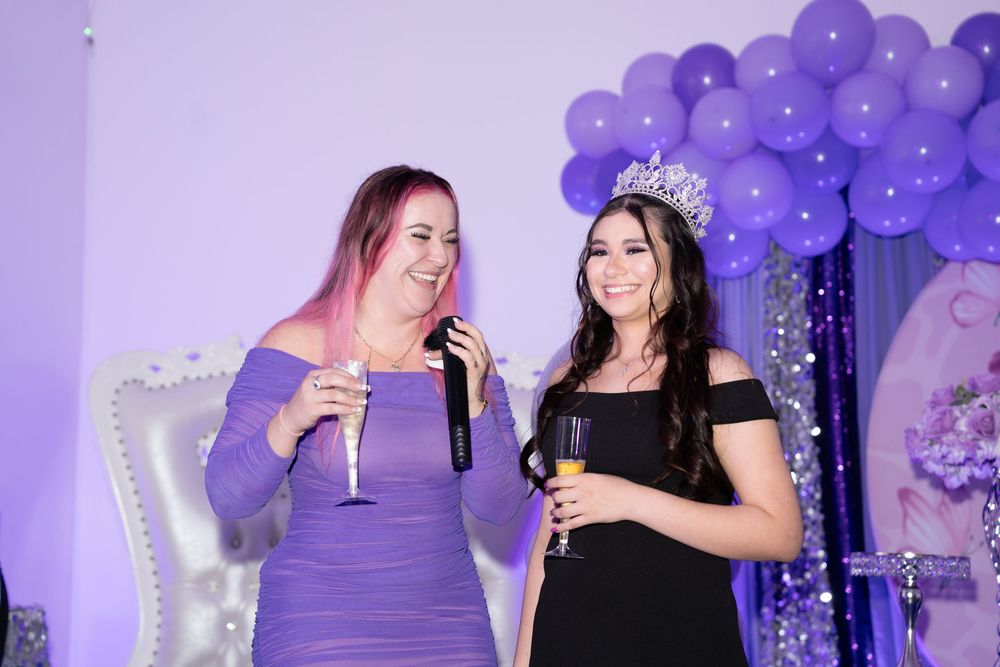 Two women in purple and black dresses celebrate with champagne under purple balloon arch, one wearing a tiara.