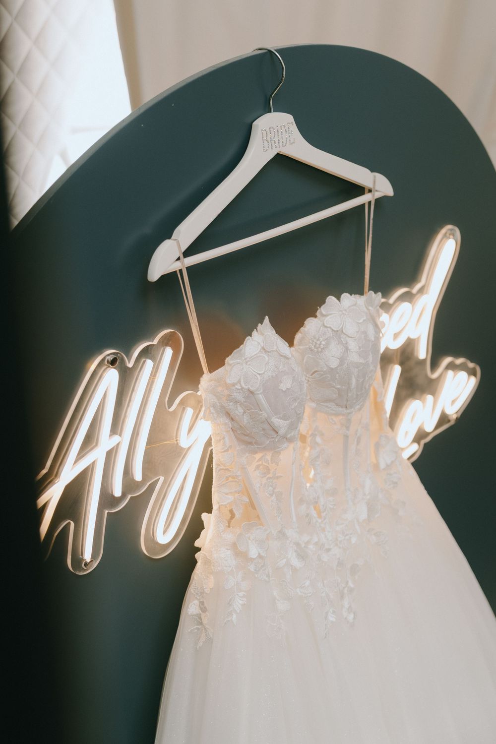 Elegant white floral wedding dress on hanger against green backdrop with glowing neon sign reading All You Need is Love.