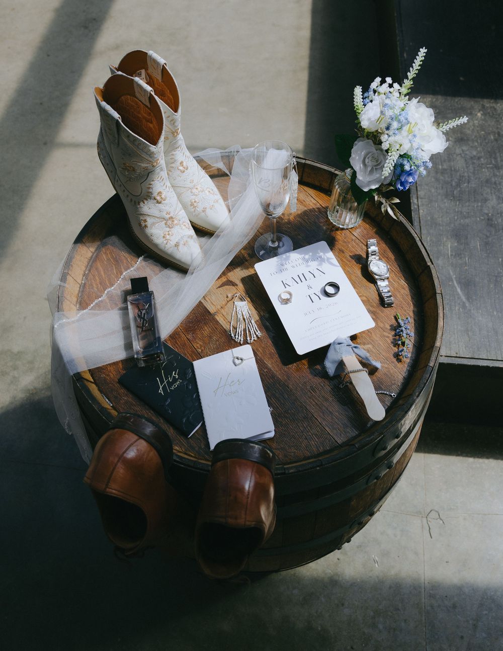 Wedding flat lay on rustic barrel featuring bridal boots, groom shoes, rings, invitations, watch, and floral bouquet.