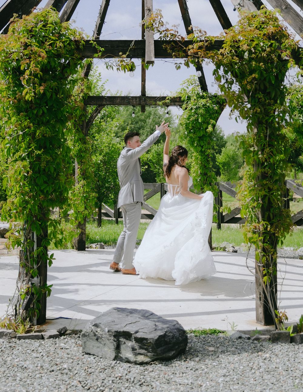 Bride and groom dancing under a lush green wooden gazebo on their wedding day in an outdoor garden setting.
