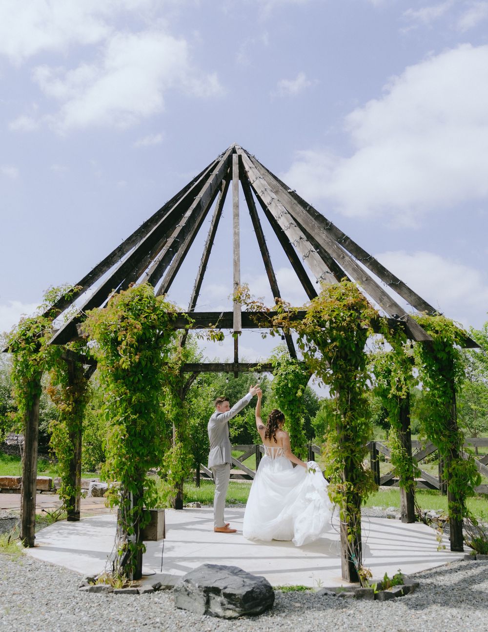 A bride and groom dance under a lush green ivy-covered wooden gazebo on their wedding day.