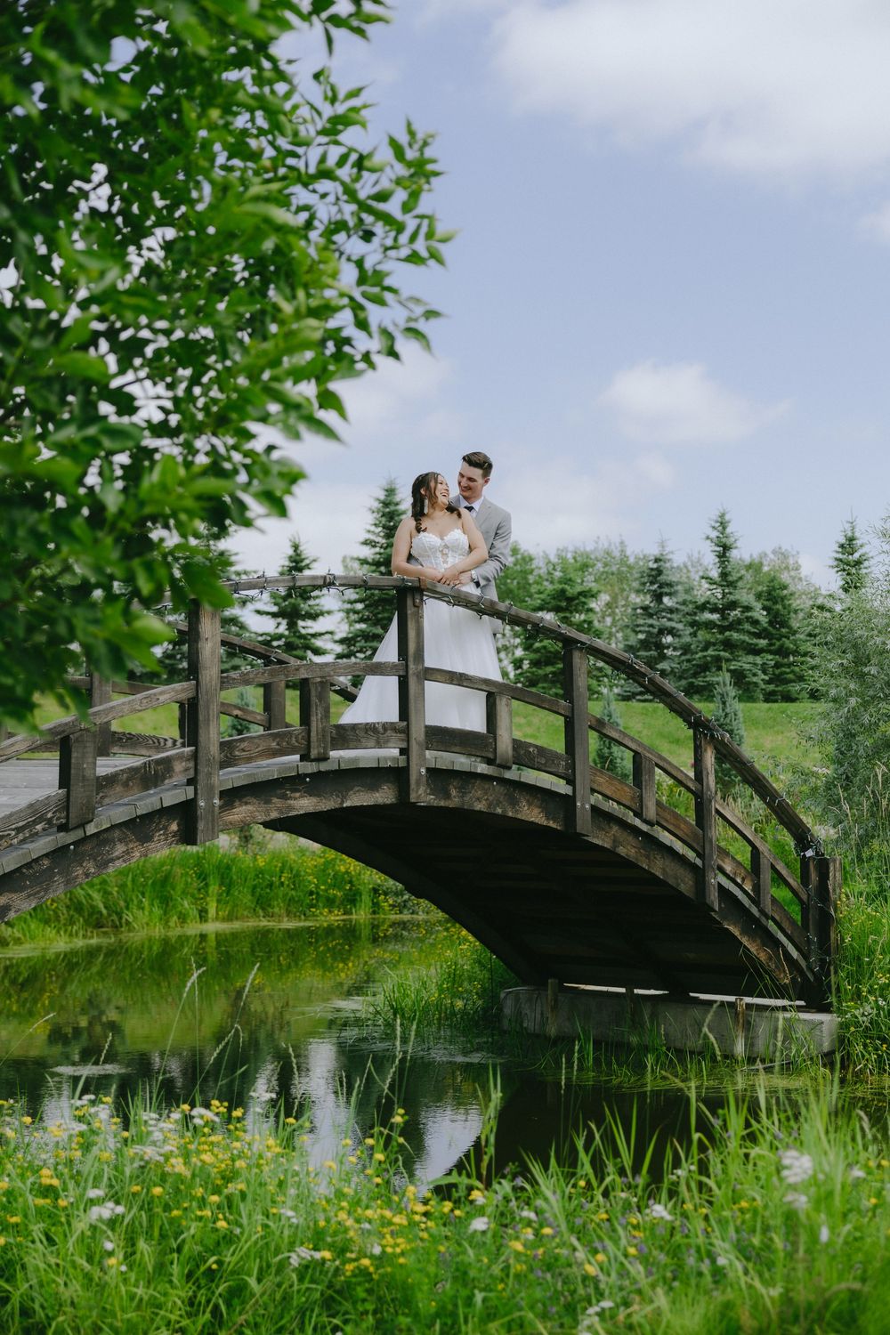 Bride and groom embrace on a rustic wooden arch bridge surrounded by lush greenery and a serene pond reflection.