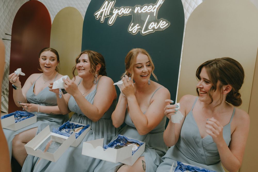 Four bridesmaids in grey dresses laughing and opening gift boxes under an All You Need Is Love neon sign.